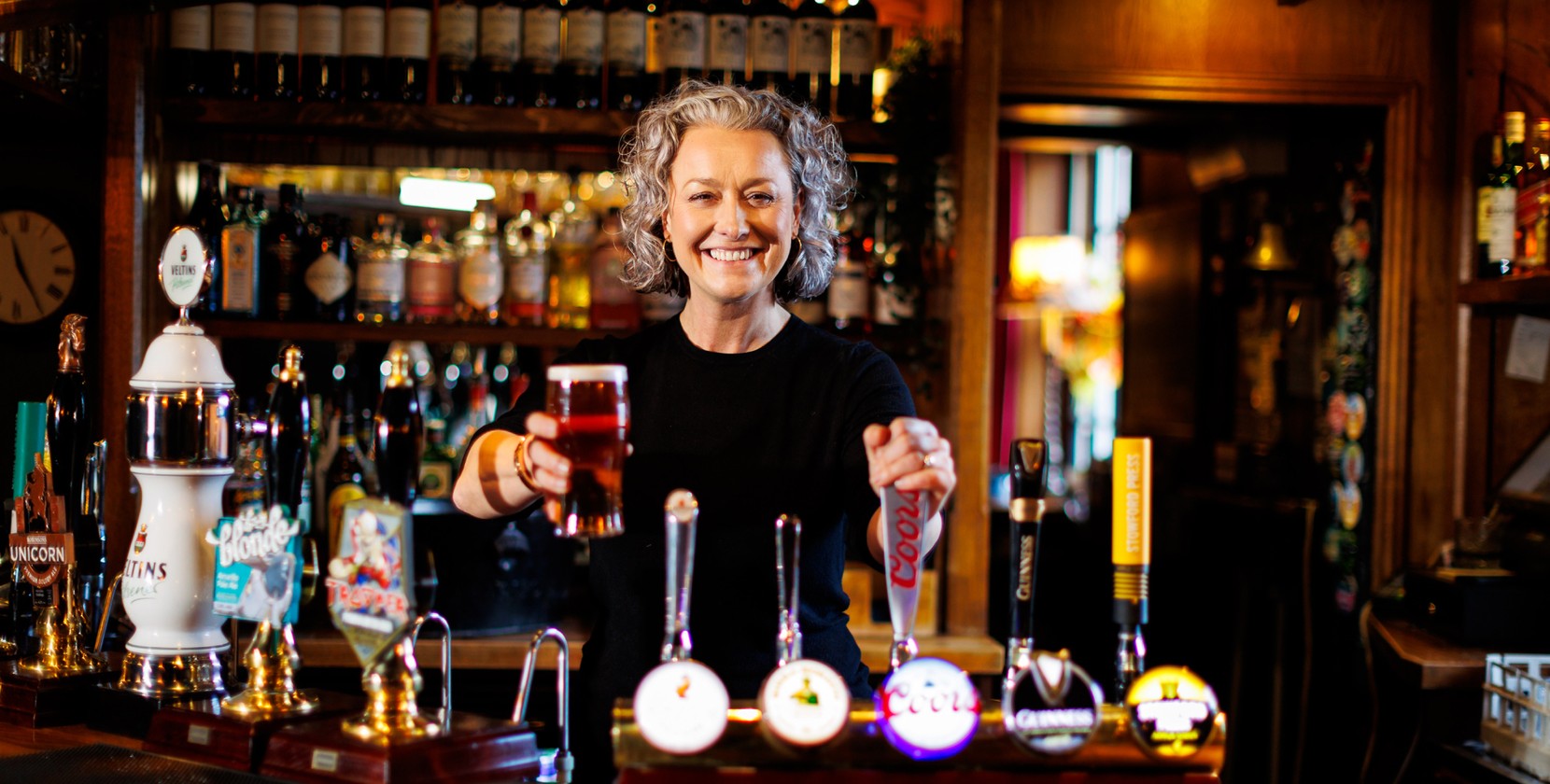 A women smiling while pulling a pint behind the bar of a Robinsons pub.