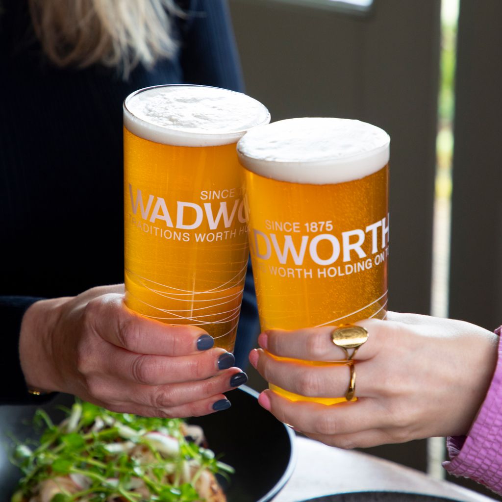 Two hands clinking pint glasses filled with golden beer, both glasses branded with the Wadworth logo and the text ‘Since 1875 – Traditions Worth Holding On To’. One hand wears a gold ring, and a plate of food with green garnish is partially visible in the foreground. 