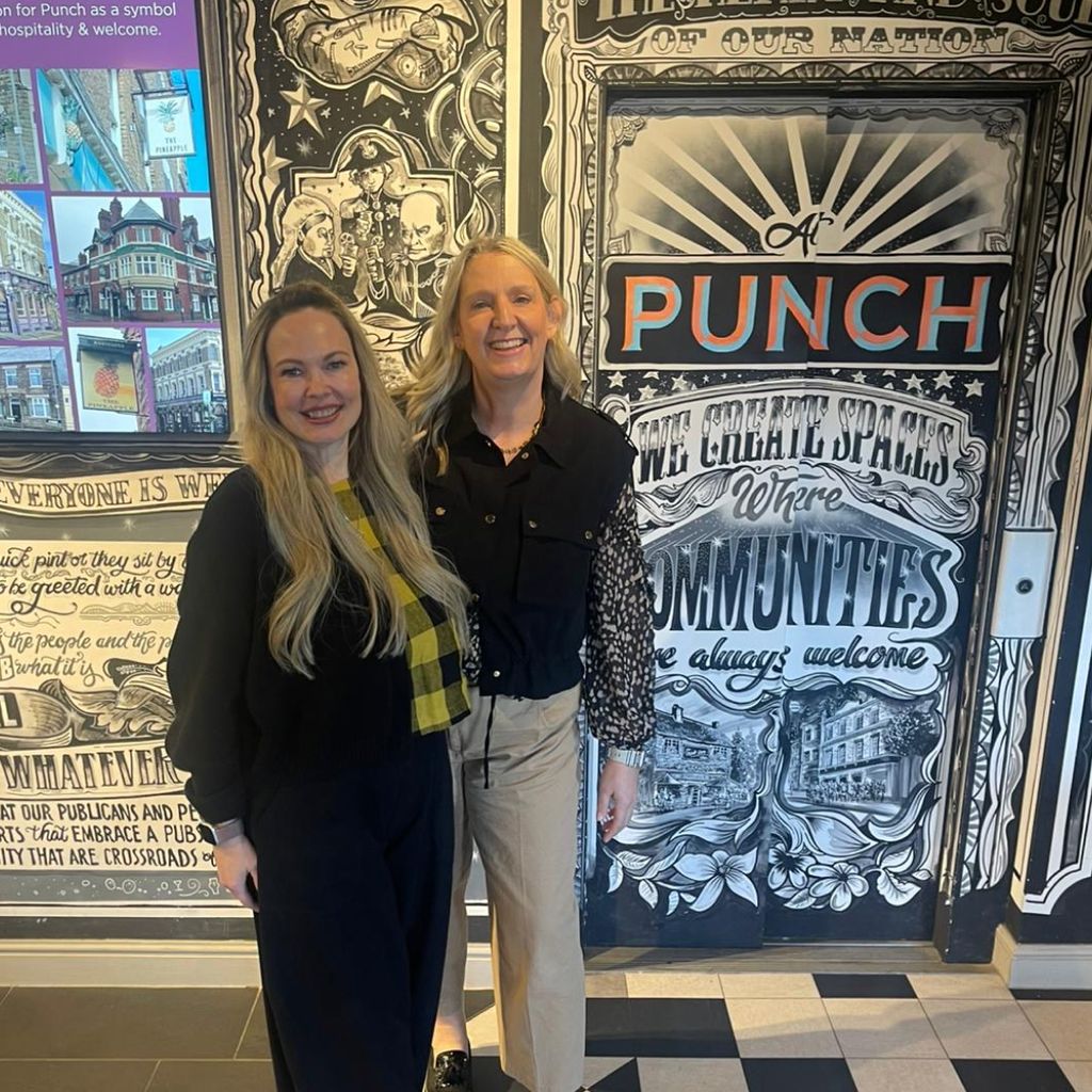 Emm and Max stand smiling side by side in front of an illustrated black, white and orange mural. The mural includes text reading "At Punch, we create spaces where communities are always welcome" alongside decorative Victorian-style illustrations and a photo collage of pub buildings.