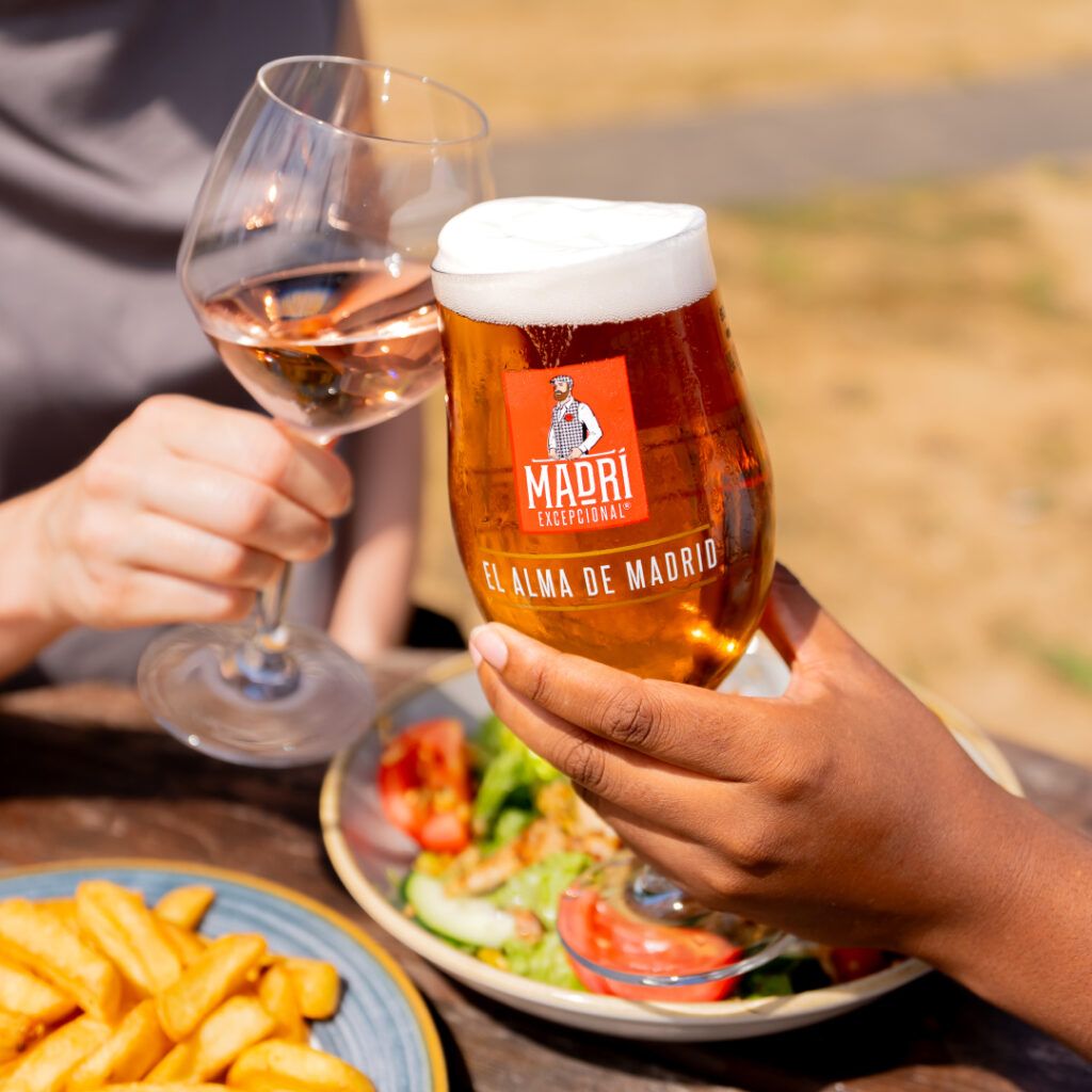 A close-up photo of two people “cheers-ing” over a wooden table with a glass of Madri and a glass of rose wine. Out of focus food can be seen in the background, including a plate of salad and a bowl of chips.
