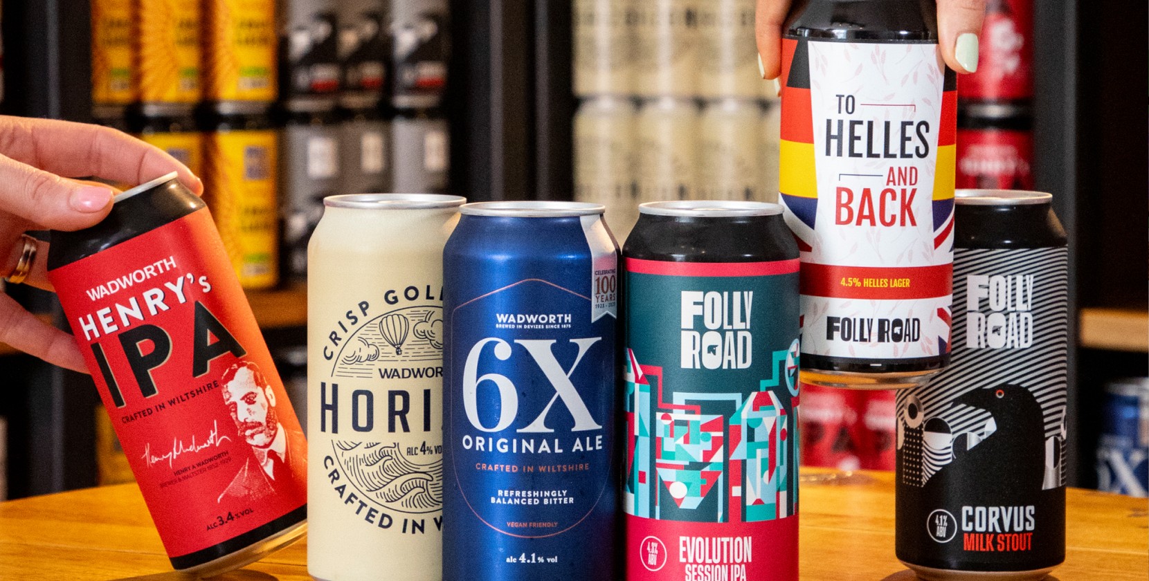 Close-up of six craft beer cans displayed on a wooden surface, featuring colourful designs and branding. Visible labels include Wadworth Henry’s IPA in red, Horizon in cream, 6X Original Ale in blue, Folly Road Evolution Session IPA in teal and red, To Helles and Back in white with bold stripes, and Corvus Milk Stout in black with a crow illustration. Background shows shelves stocked with more cans. 