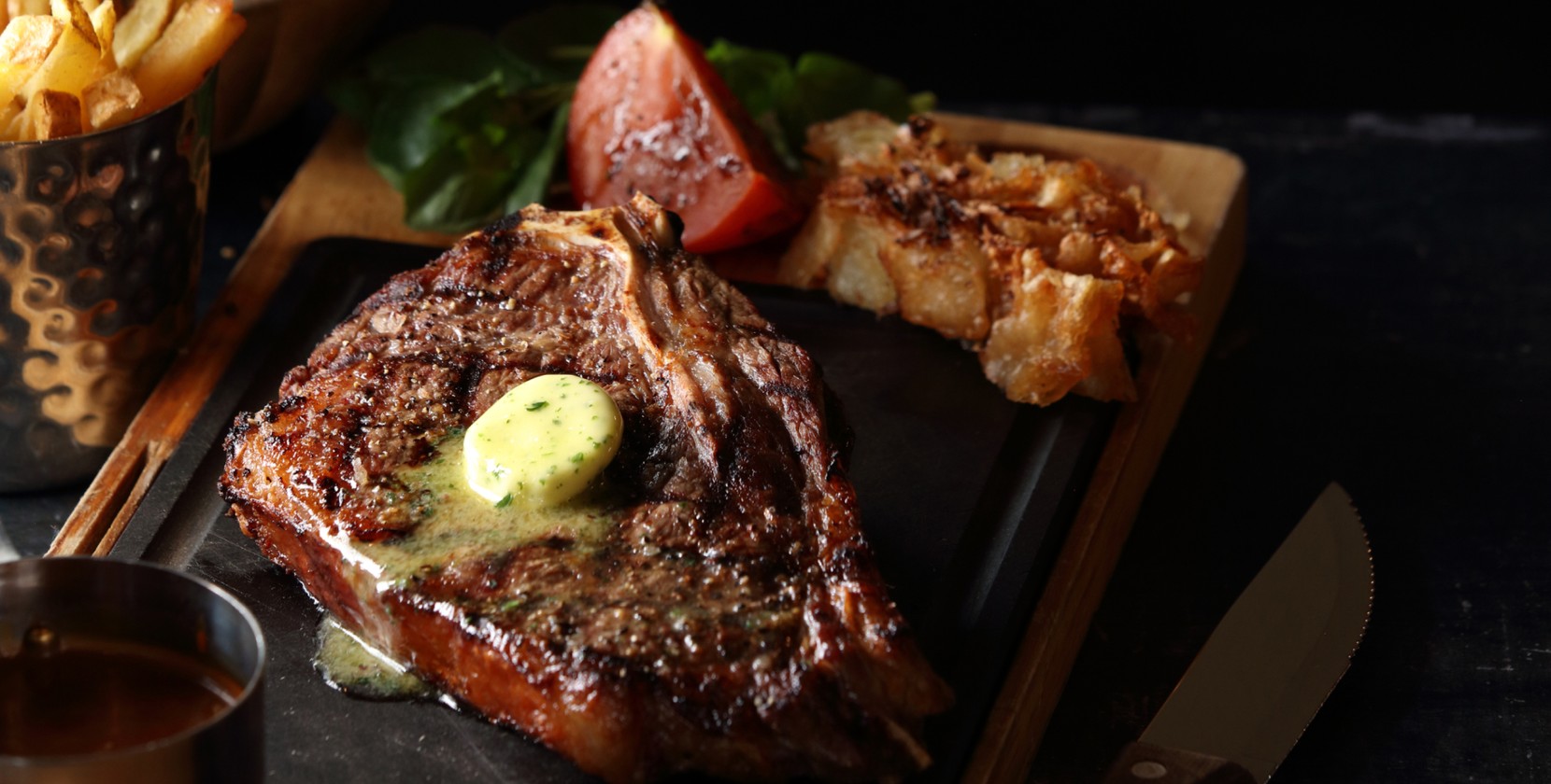 A close-up photograph of a steak on a rustic serving plate. The steak has butter gently melting on the top side. Other menu items can be seen in the background, including a tomato and greens.