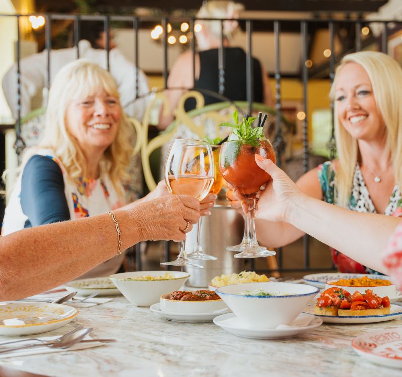 A group of women sit around a table in the Italian restaurant, Pesto, raising their wine glasses in a cheers motion. Various plates of food can be seen on the table along with other people in the background.