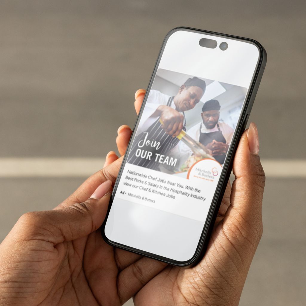 Someone holding an iPhone; on its screen is an example of a HR recruitment ad for Mitchells &amp; Butlers. 