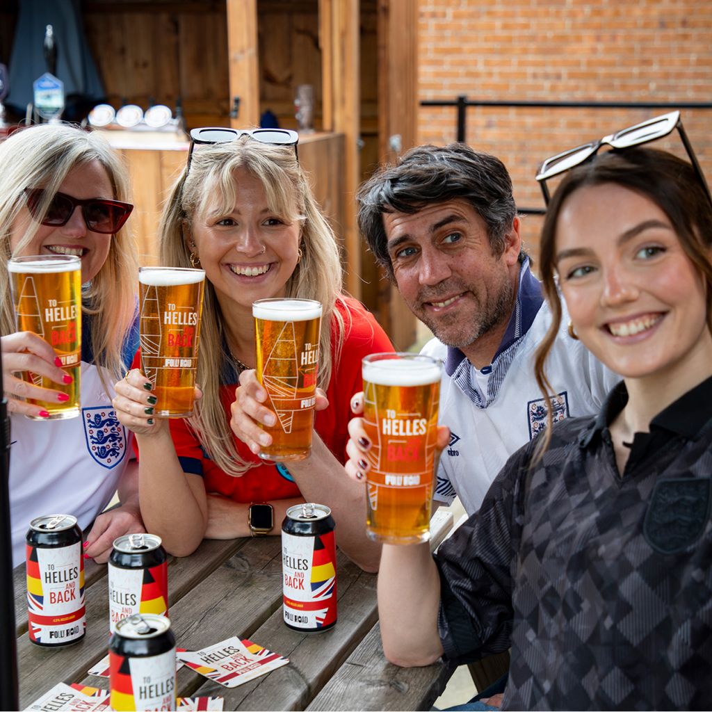 Group of people sitting at a wooden outdoor table holding pints of beer branded ‘To Helles and Back’. Several matching beer cans and promotional leaflets are placed on the table. 