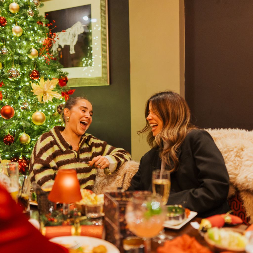 Two people laugh heartily on a sofa in front of a large, decorated Christmas tree.