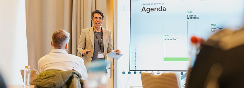 Presenter speaking to a small audience beside a screen showing an agenda.