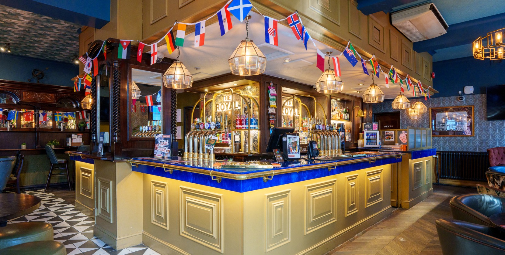 A classic British pub bar during the World Cup; around the edge of the bar, all the countries' flags are hung. Blue accents and patterns catch the eye.