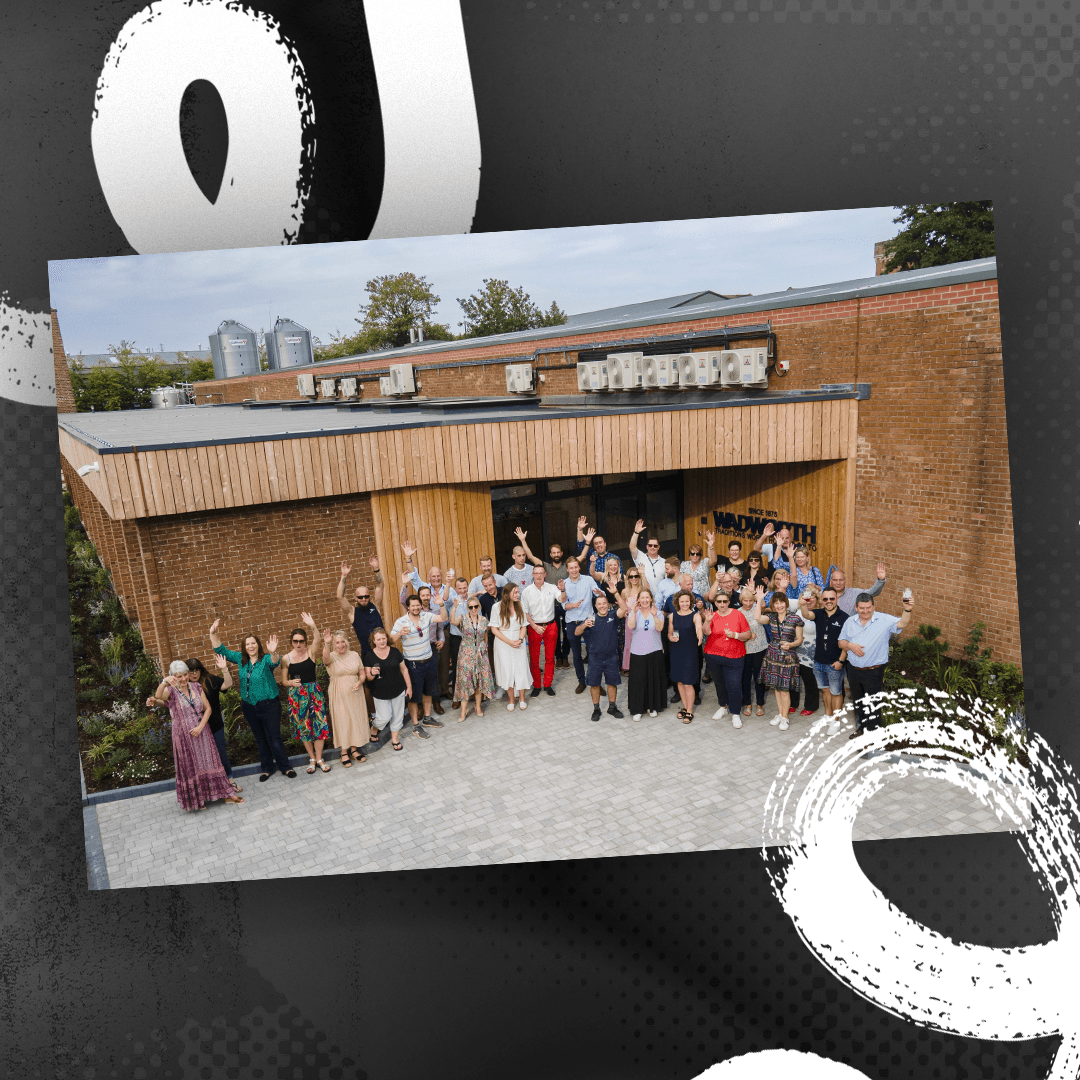Large group of people standing outside a modern brick building with wooden cladding, posing together and raising their arms in celebration. The building has a sign reading ‘WADWORTH’ near the entrance, and the background shows trees and industrial silos. The photo is framed with abstract white brushstroke graphics on a dark textured backdrop. 