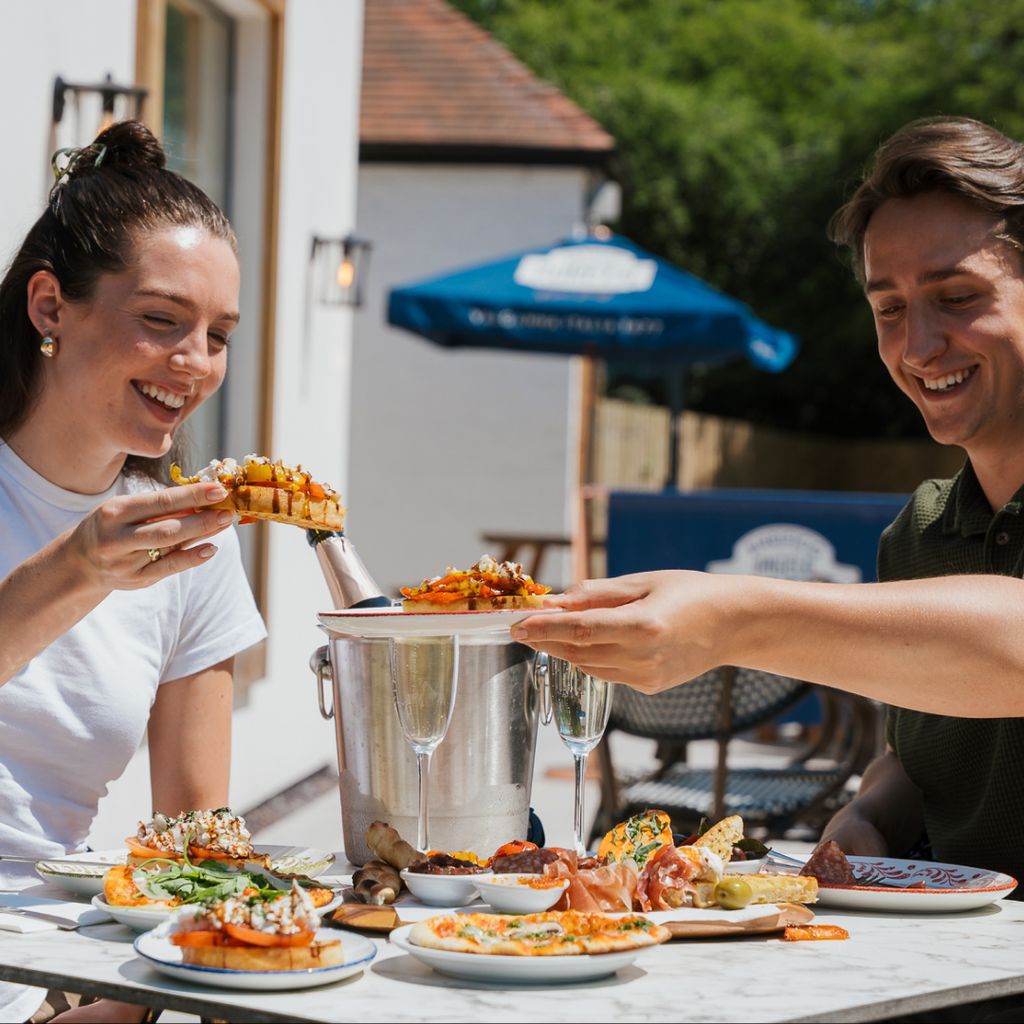 Two people sit at an outside table enjoying Italian food at Pesto. On the dining table a champagne ice bucket, various plates of food, and two champagne flutes are visible.