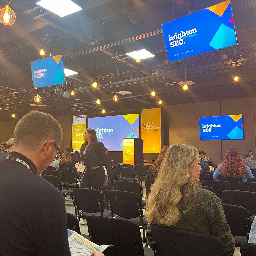 The interiors of BrightonSEO, with people taking their seats. At the front, multiple screens with the event logo and design.