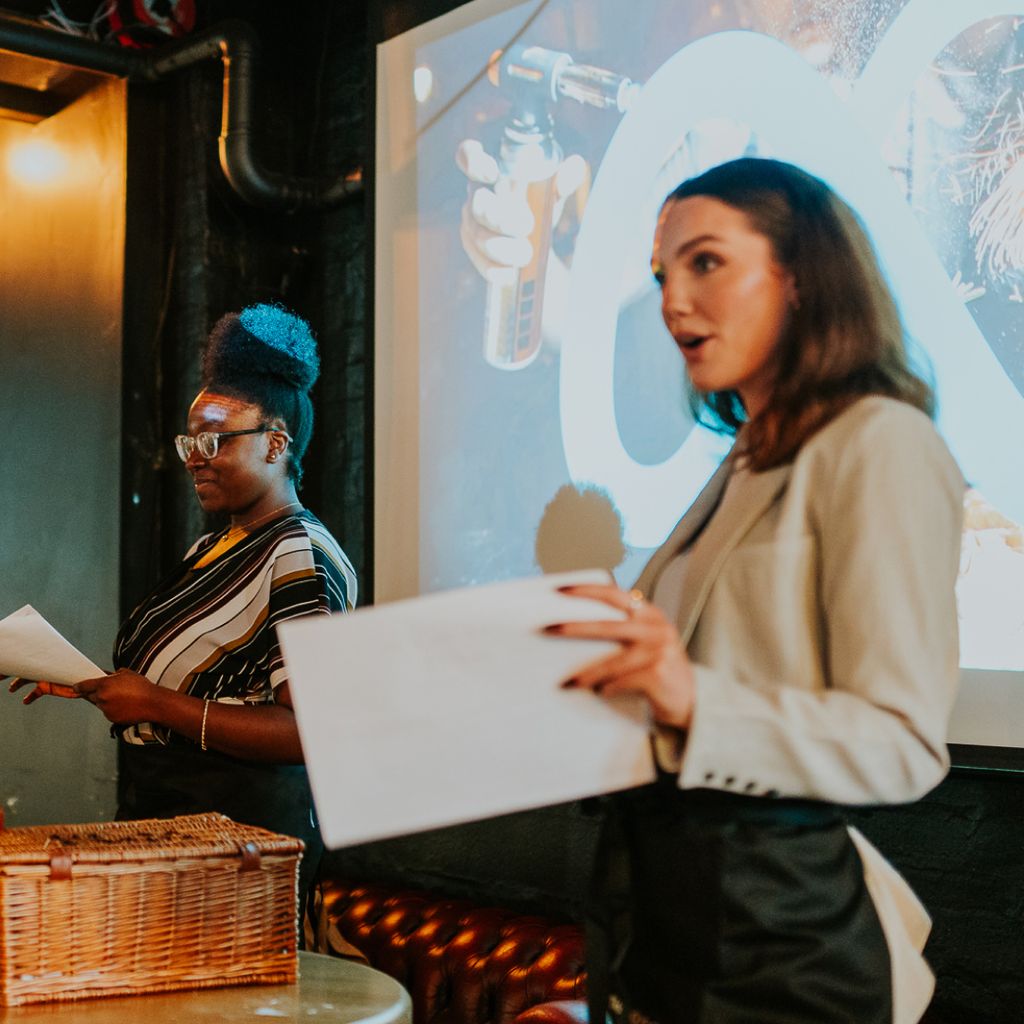 Beatrice wears a beige blazer and speaks while holding papers, standing in front of a projected screen. Brithney wears glasses and stands beside Beatrice, holding notes, in a dimly lit venue with a wicker hamper on a table in the foreground.