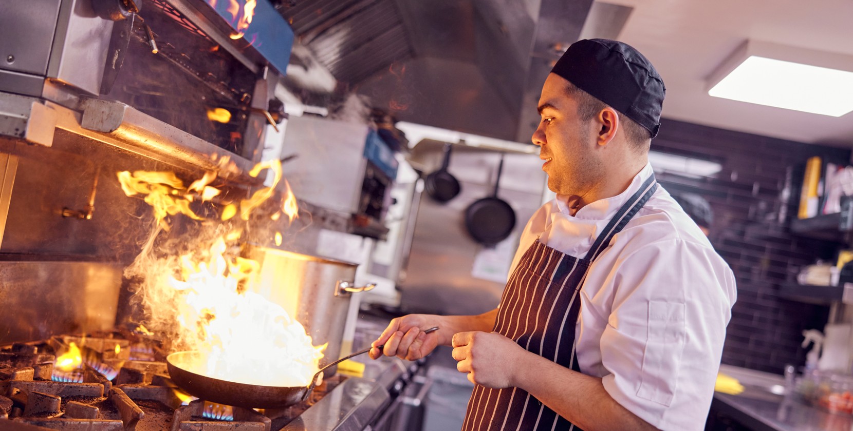 A chef wearing a white uniform and black apron cooking in a commercial kitchen, holding a frying pan over an open flame with visible flames rising from the pan. Stainless steel appliances and utensils hang in the background.
