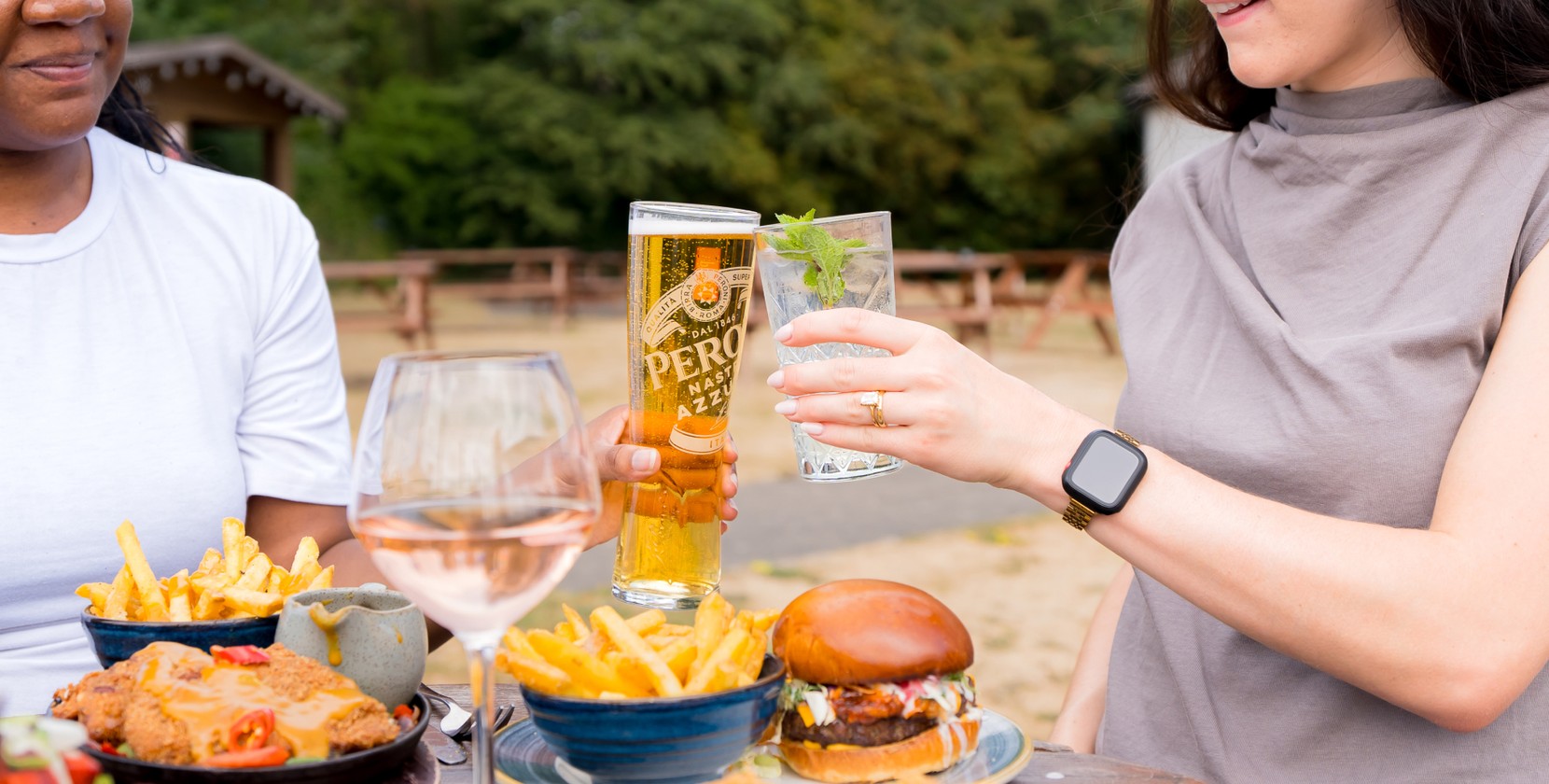 A photo of two smiling people sitting at a wooden picnic bench in a large UK beer garden. There is a selection of food and drink on the table, including a loaded burger with fries. One person holds a glass of Peroni, and the other holds a soft drink, and they are clinking the glasses together in a cheers motion.