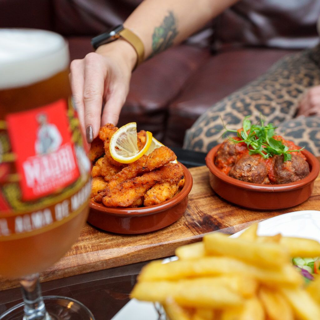 A close-up photo of food on a table in a pub. A hand is reaching into frame to pick up some of the food. To the left of the frame is an out of focus glass of Madri and a bowl of chips.